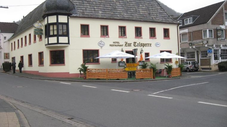 Ein charmantes Restaurant und Hotel namens "Zur Talsperre" an einer Straßenecke. Es hat eine einladende Terrasse mit Sonnenschirmen und ist von einer ruhigen Straße umgeben.