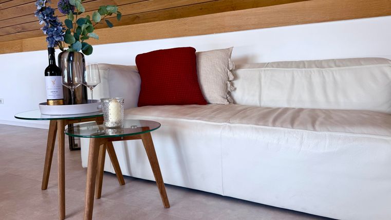 Eine gemütliche Couch mit einem roten Kissen und einer weichen Decke. Neben der Couch steht ein stilvoller Tisch mit Blumen, einer Flasche Wein und einem Glas.