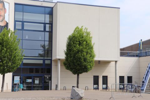 Ein modernes Gebäude mit großen Fenstern und einem klaren Himmel. Vor dem Gebäude stehen zwei kleine Bäume und einige Fahrräder sind abgestellt.