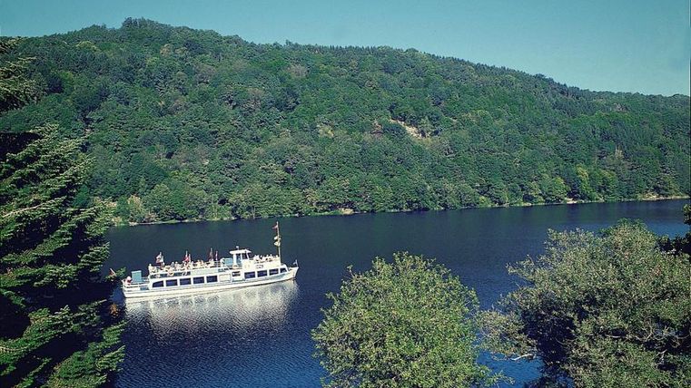 Ein Boot fährt auf einem ruhigen See, umgeben von grünen Bäumen und Hügeln. Die Landschaft ist malerisch und einladend.