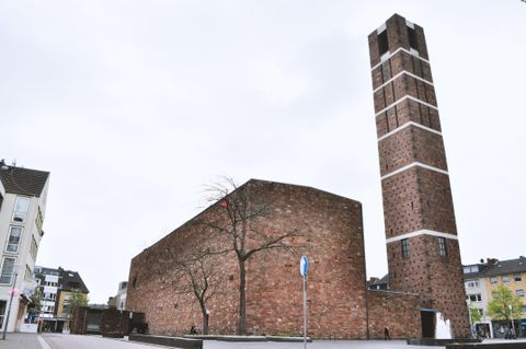 Ein markantes Gebäude aus roten Ziegeln mit einem hohen Schornstein. Die Umgebung ist städtisch mit grauem Himmel und vereinzelten Bäumen.