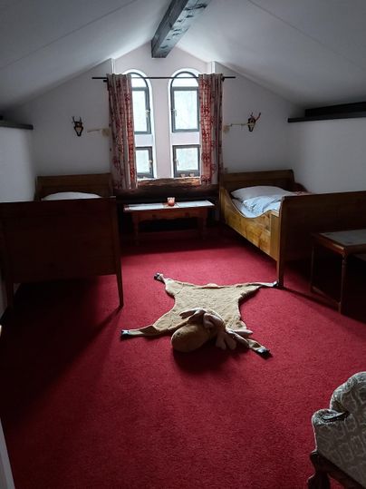 Ein gemütliches Zimmer mit zwei Betten und einem roten Teppich. An der Wand hängen Fenster mit Vorhängen und im Raum liegt ein Tierfell.