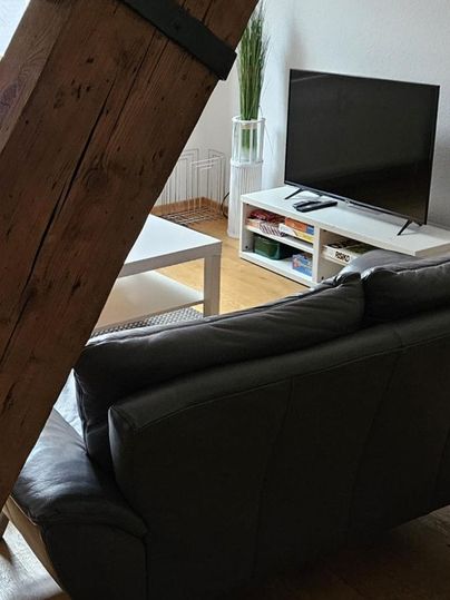 Ein gemütliches Wohnzimmer mit einem grauen Sofa und einem Fernseher. Im Hintergrund steht ein weißer Couchtisch und eine Pflanze.