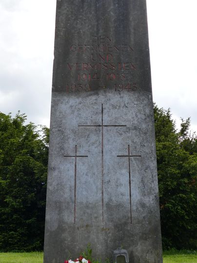 Ein Gedenkstein mit drei Kreuzsymbolen und einer Inschrift. Vor dem Stein sind Blumen und eine kleine Laterne platziert.