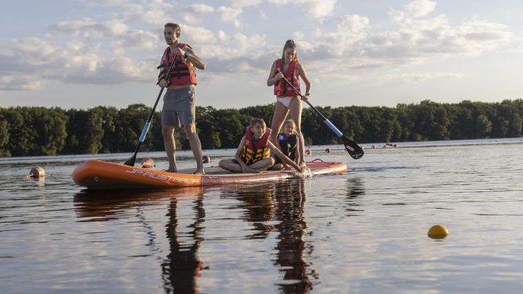 Eine Gruppe von vier Personen paddelt auf einem Stand-up-Paddleboard auf einem ruhigen See. Sie tragen Rettungswesten und genießen das schöne Wetter.