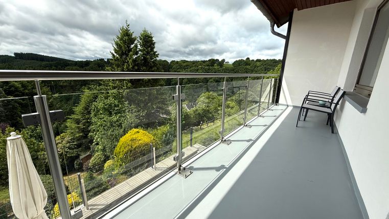Ein Balkon mit einer Glasgeländer und einem Blick auf die grüne Landschaft. Es gibt zwei Stühle und einen Sonnenschirm auf der linken Seite.