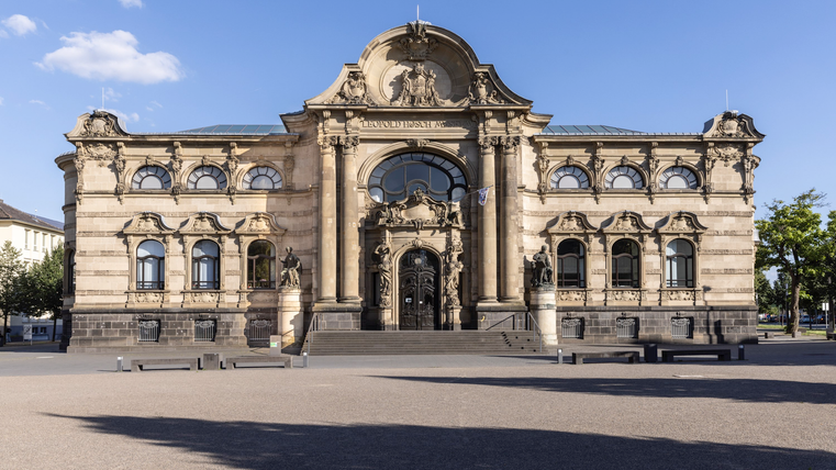 Eine Hand hält ein gebogenes Blatt Papier. Der Hintergrund enthält Informationen über eine Ausstellung im Leopold-Hoesch-Museum.