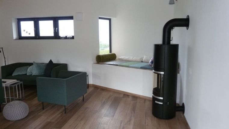 Ein moderner Wohnraum mit gemütlicher grüner Couch und einem Holzofen in der Ecke. Helle Wände und große Fenster sorgen für viel Tageslicht.