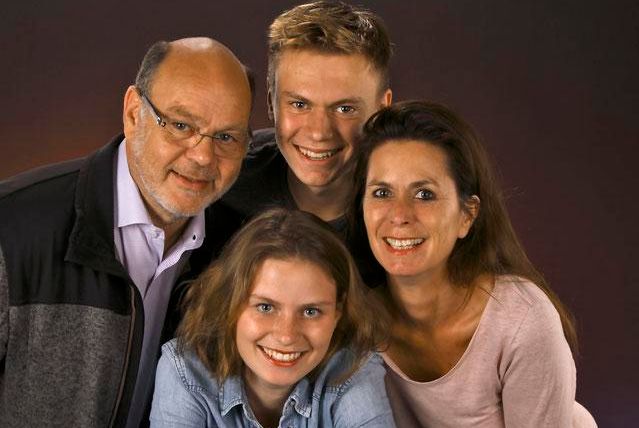Eine fröhliche Familie steht zusammen und lächelt in die Kamera. Sie bestehen aus zwei Erwachsenen und zwei Jugendlichen.