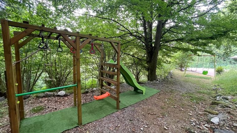 Ein Spielplatz im Freien mit einer Rutsche und Klettergerüst. Umgeben von Bäumen und grünem Gelände.