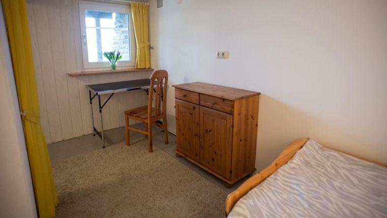 Ein einfaches Zimmer mit einem Bett, einem Holzschreibtisch und einer Kommode. Gelbe Vorhänge lassen Tageslicht herein.