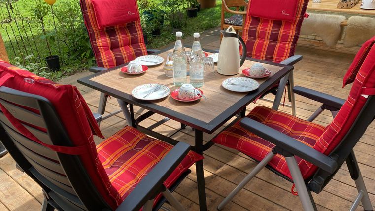 Ein schön gedeckter Tisch auf einer Terrasse mit roten Stühlen. Auf dem Tisch stehen Getränke und Teller für eine gemütliche Runde.