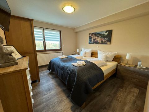 Ein gemütliches Schlafzimmer mit einem großen Bett und modernen Möbeln. Die Wände sind in sanften Farben gehalten und große Fenster lassen viel Licht herein.