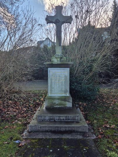 Ein alten Grabstein mit einem Kreuz steht in einem bewachsenen Bereich. Umgeben von trockenen Zweigen und Gras ist die Szene ruhig und besinnlich.