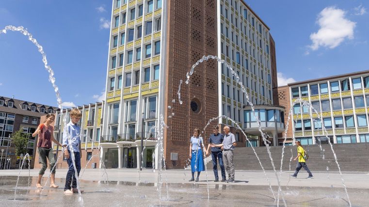 Ein modernes Gebäude mit einer Wasserspiele-Anlage im Vordergrund. Menschen genießen die frische Brise an einem sonnigen Tag.