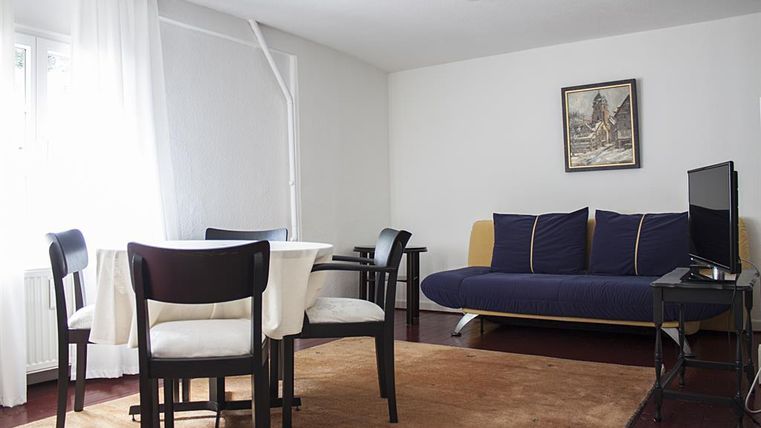 Ein helles Wohnzimmer mit einem Tisch und Stühlen. Es gibt ein Sofa und einen Fernseher an der Wand.