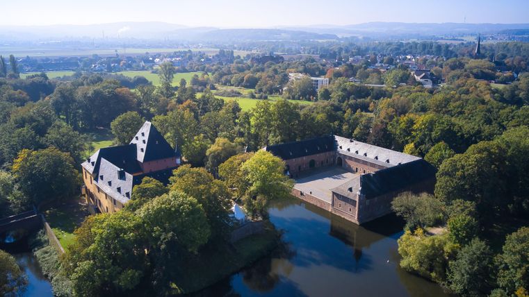 Eine malerische Ansicht eines Schlosses umgeben von Bäumen und einem ruhigen Wasser. Im Hintergrund erstreckt sich eine sanfte Landschaft unter einem klaren Himmel.