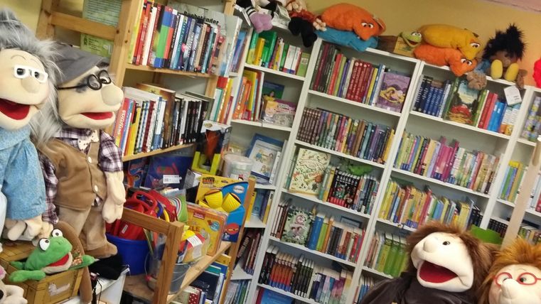 Ein gemütlicher Raum voller Bücherregale und Handpuppen. Die bunten Puppen sitzen neben einer kleinen Leseecke und schaffen eine einladende Atmosphäre.