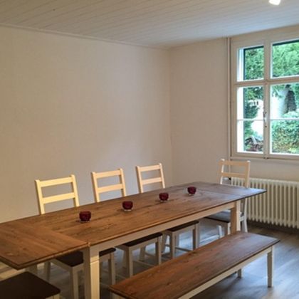 Ein helles Esszimmer mit einem Holztablett und weißen Stühlen. Durch das Fenster ist eine grüne Aussicht zu sehen.