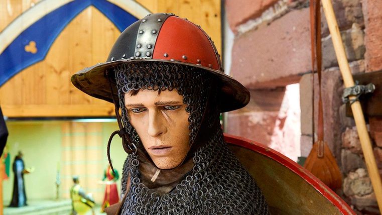 Eine realistische Figur eines Ritters mit einem Kettenhemd und einem auffälligen Helm. Im Hintergrund sind weitere historische Objekte sichtbar.