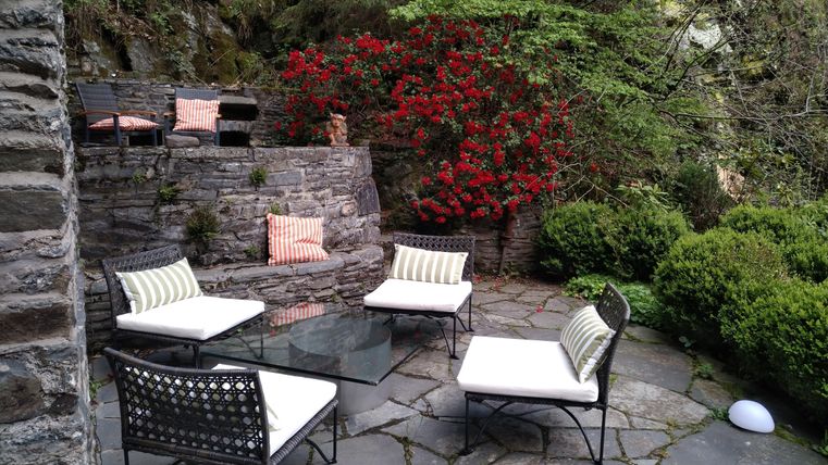 Eine gemütliche Terrasse mit modernen Möbeln und einem Glastisch. Im Hintergrund blühen rote Pflanzen und der Garten ist grün und einladend.