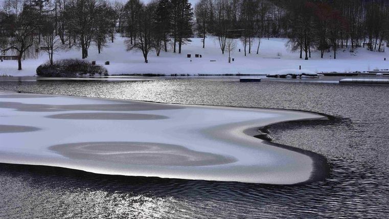 Ein gefrorener See mit schneebedecktem Ufer und vereistem Wasser. Im Hintergrund sind kahle Bäume und eine ruhige Winterlandschaft zu sehen.