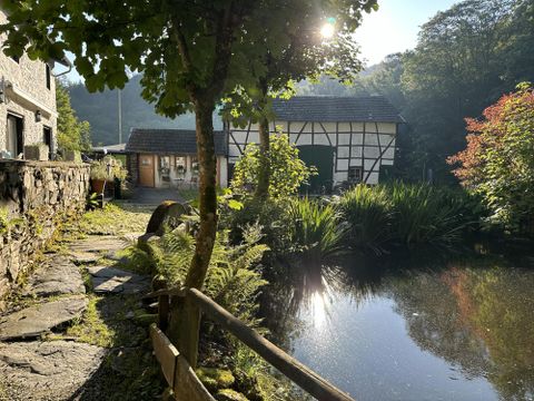Ein malerischer Teich umgeben von üppigem Grün und Bäumen. Im Hintergrund ist ein traditionelles Haus mit Fachwerkstil zu sehen.