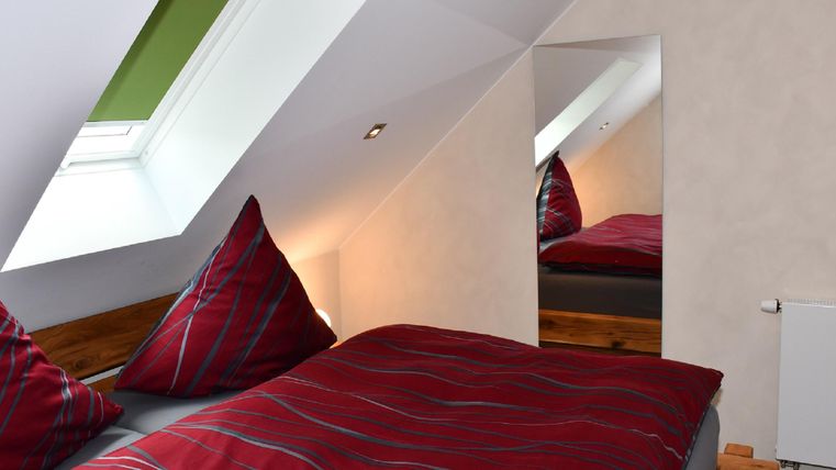 Ein gemütliches Dachzimmer mit einem großen Bett und bunten Kissen. Ein Spiegel und ein Dachfenster sorgen für zusätzliches Licht.