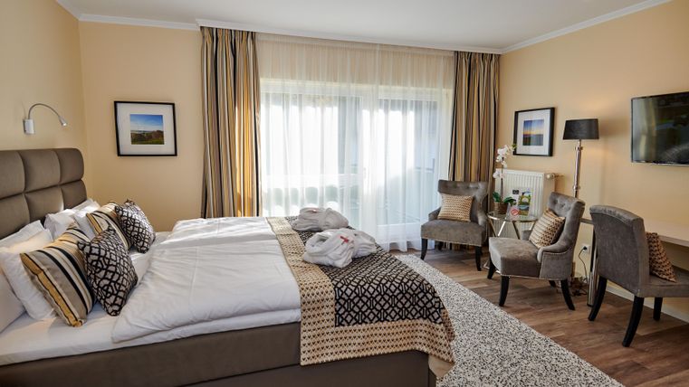 Ein elegantes Hotelzimmer mit einem Doppelbett und bequemen Sesseln. Große Fenster sorgen für viel Tageslicht und eine gemütliche Atmosphäre.