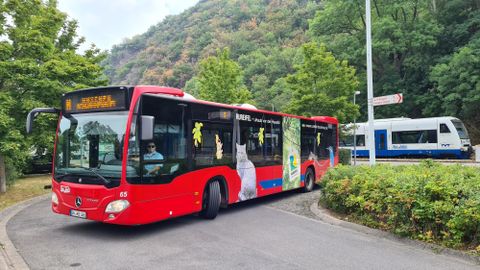 Ein roter Bus fährt eine Kurve, während ein Zug in der Nähe hält. Im Hintergrund sind grüne Bäume und ein bergiger Hang zu sehen.