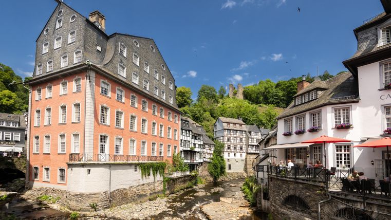 Eine malerische Stadt mit historischen Gebäuden an der Rur. Im Hintergrund sind grüne Hügel und ein blauer Himmel zu sehen.