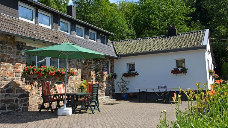Ein gemütliches Haus mit einem gedeckten Außentisch und einer grünen Markise. Den Innenhof umgeben Blumen und Bäume, und der Himmel ist blau.