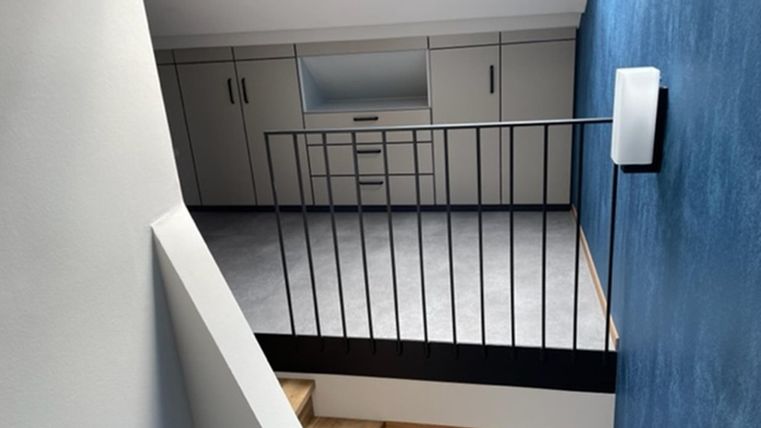 Ein moderner Raum mit einer blauen Wand und einer Holztreppe. Auf der anderen Seite gibt es eine helle Wand mit Schränken und einem Fenster.