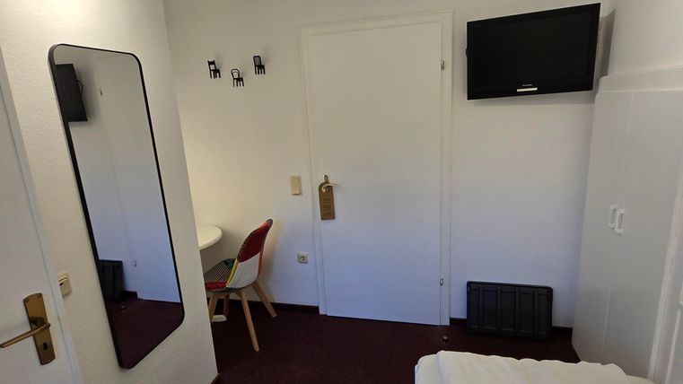 Ein einfaches Hotelzimmer mit einem Bett, einem Tisch und einem Fernseher an der Wand. Es gibt einen großen Spiegel und helles Licht durch die Decke.