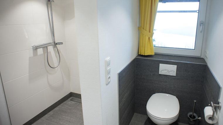 Ein modernes Badezimmer mit einer separaten Dusche und einer Toilette. Helle Wände und ein Fenster mit gelben Vorhängen sorgen für eine freundliche Atmosphäre.