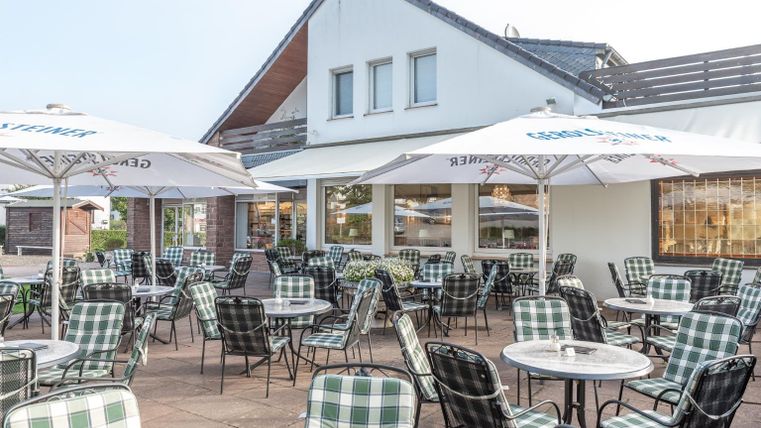 Eine Terrasse mit vielen Tischen und Stühlen, umgeben von Sonnenschirmen. Im Hintergrund ist ein modernes Gebäude zu sehen.
