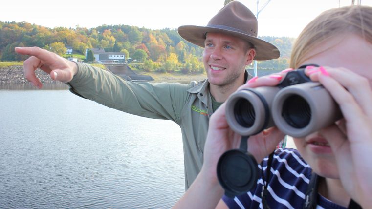 Ein Mann zeigt auf den See, während ein Kind mit einem Fernglas beobachtet. Im Hintergrund sind Bäume mit herbstlichen Farben zu sehen.