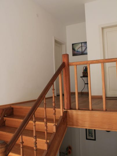 Ein heller Flur mit einer Holztreppe und einem Geländer. Im Hintergrund sind mehrere Türen und ein Tisch zu sehen.