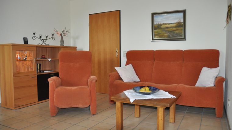 Ein gemütliches Wohnzimmer mit einem orangefarbenen Sofa und einem Holztisch. Die Wände sind hell und es gibt ein Bild sowie eine kleine Vitrine.