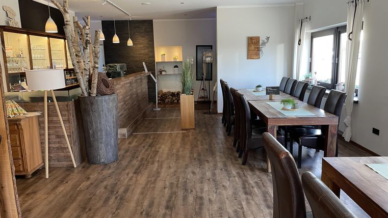 Ein modernes Restaurant mit Holzfußboden und großen Fenstern. Tische sind für Gäste eingedeckt und eine einladende Atmosphäre ist sichtbar.