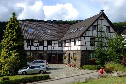 Ein schönes Fachwerkhaus mit einem gepflegten Garten. Vor dem Haus stehen parkende Autos und eine Person sitzt auf der Bank.