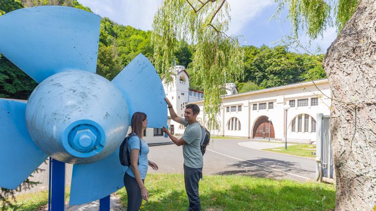 Ein großes, blaues Schaufelrad steht im Vordergrund, während zwei Personen davor stehen und miteinander sprechen. Im Hintergrund sind historische Gebäude und viel Grün zu sehen.