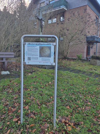 Ein Informationsschild steht in einem Garten mit herbstlichen Blättern. Im Hintergrund ist ein Gebäude und ein Kreuz sichtbar.