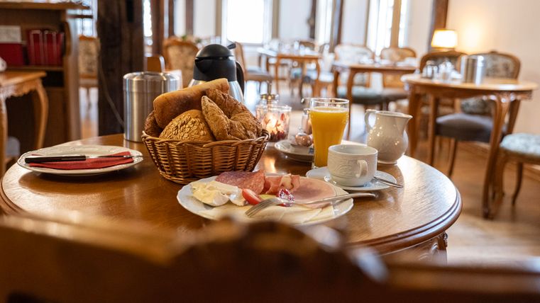 Ein gedeckter Tisch mit frischen Brötchen, Aufschnitt und einem Glas Orangensaft. Im Hintergrund sind gemütliche Stühle und ein helles Ambiente zu sehen.