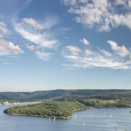 Eine malerische Landschaft mit einem ruhigen See und sanften Hügeln. Segelboote gleiten über das Wasser unter einem klaren blauen Himmel.