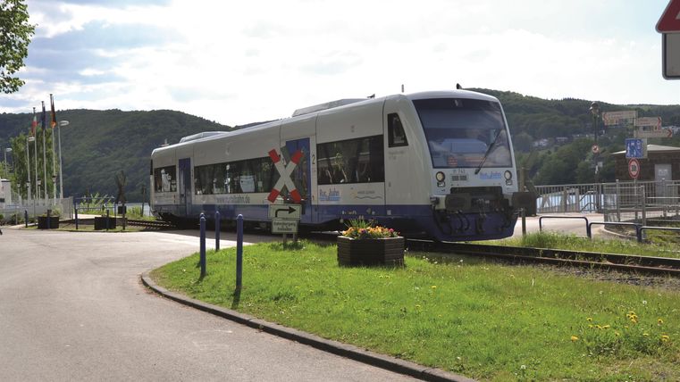 Ein moderner Zug fährt an einer Haltestelle vorbei. Die Umgebung ist grün und ländlich, mit sanften Hügeln im Hintergrund.