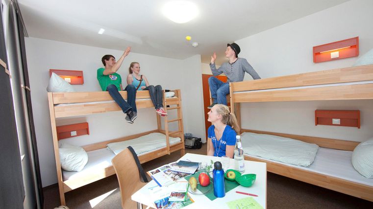 Eine lebhafte Jugendherberge mit zwei Etagenbetten und fröhlichen jungen Leuten. Sie spielen und unterhalten sich, während auf dem Tisch Snacks und Getränke liegen.