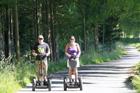 Zwei Personen fahren auf Segways entlang eines Waldwegs. Die Umgebung ist grün und sonnig.