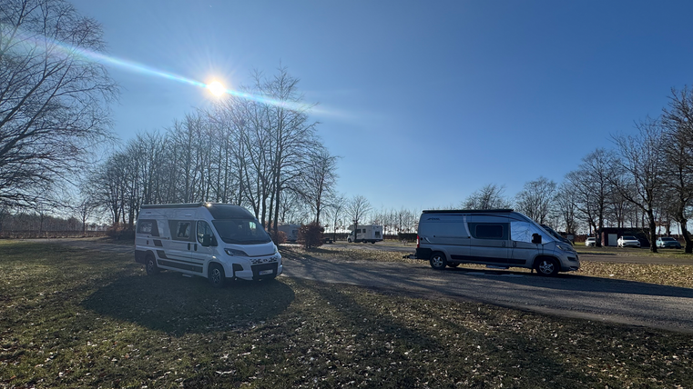 Zwei Wohnmobile stehen auf einem Platz mit Bäumen im Hintergrund. Der Himmel ist klar und sonnig.