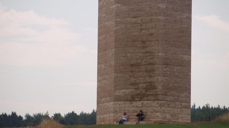 Ein hoher, rechteckiger Betonbau steht auf einem grünen Hügel. Zwei Personen sitzen davor und genießen die Aussicht.